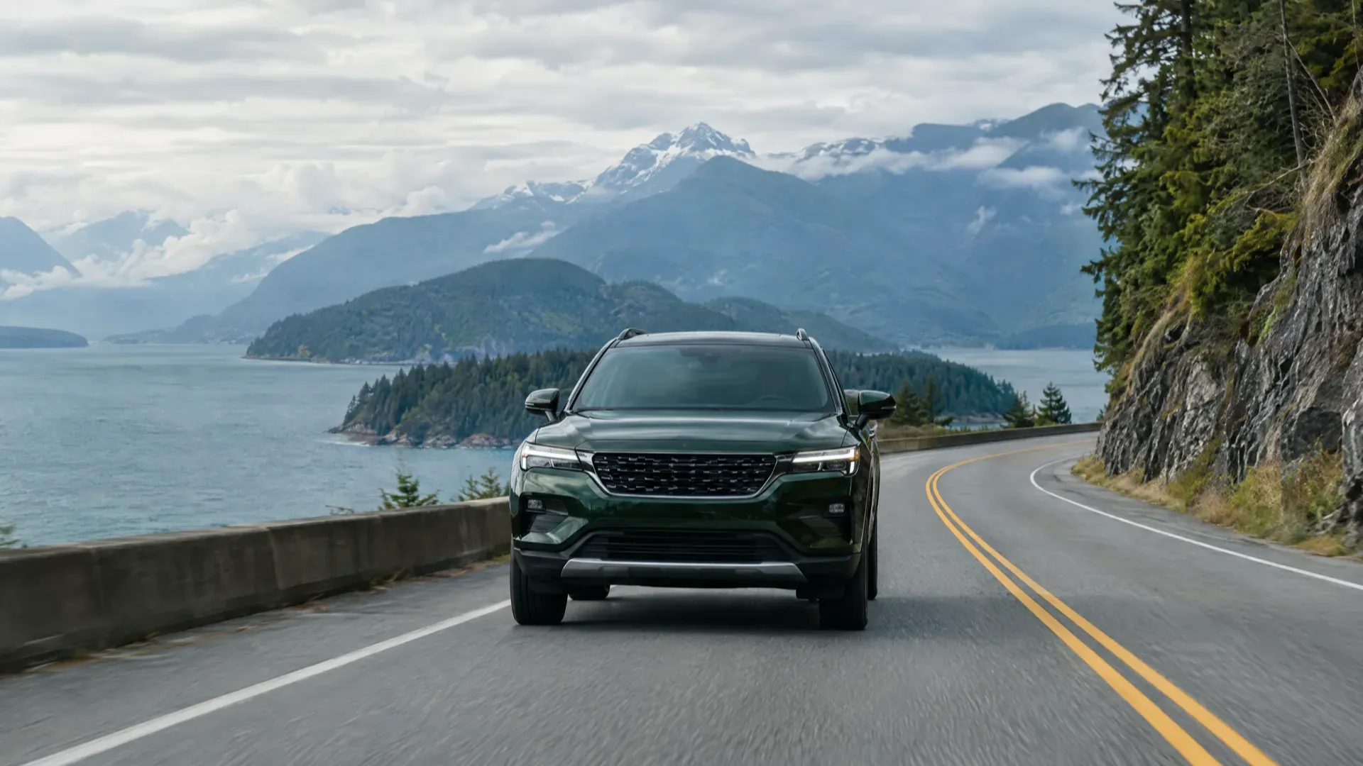 Dark green SUV on a winding coastal highway beside an ocean inlet and snow-capped mountains in British Columbia