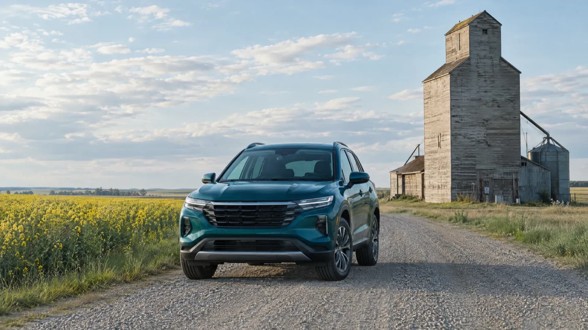 Teal crossover on a gravel road beside a canola field and a weathered wooden grain elevator on the prairies